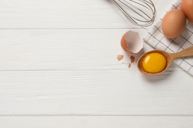 Raw chicken eggs, shell with yolk and whisk on white wooden table, flat lay. Space for text