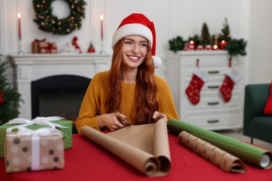 Beautiful young woman in Santa hat cutting wrapping paper at table. Decorating Christmas present