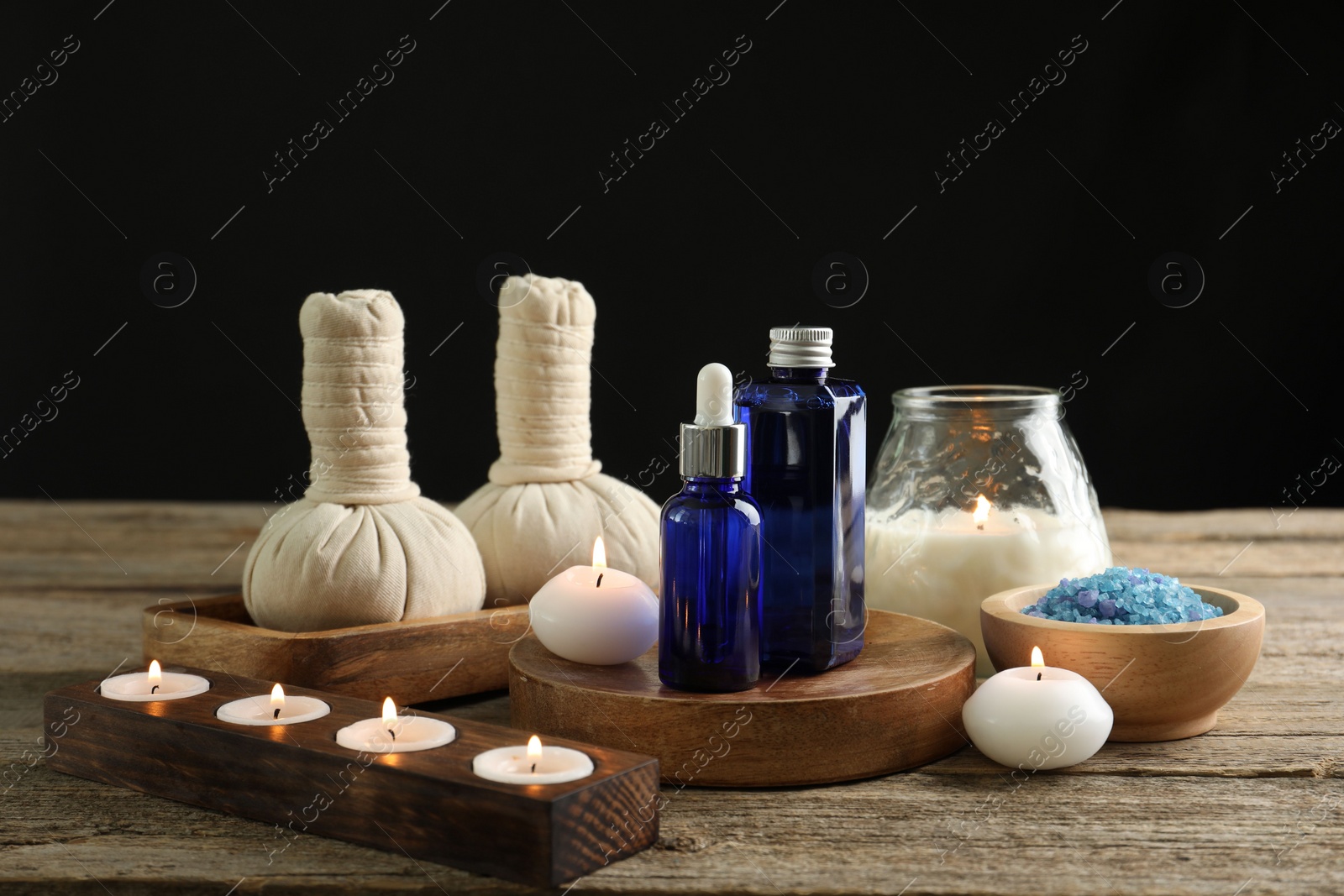 Photo of Spa composition. Cosmetic products, herbal bags, burning candles and sea salt on wooden table against black background