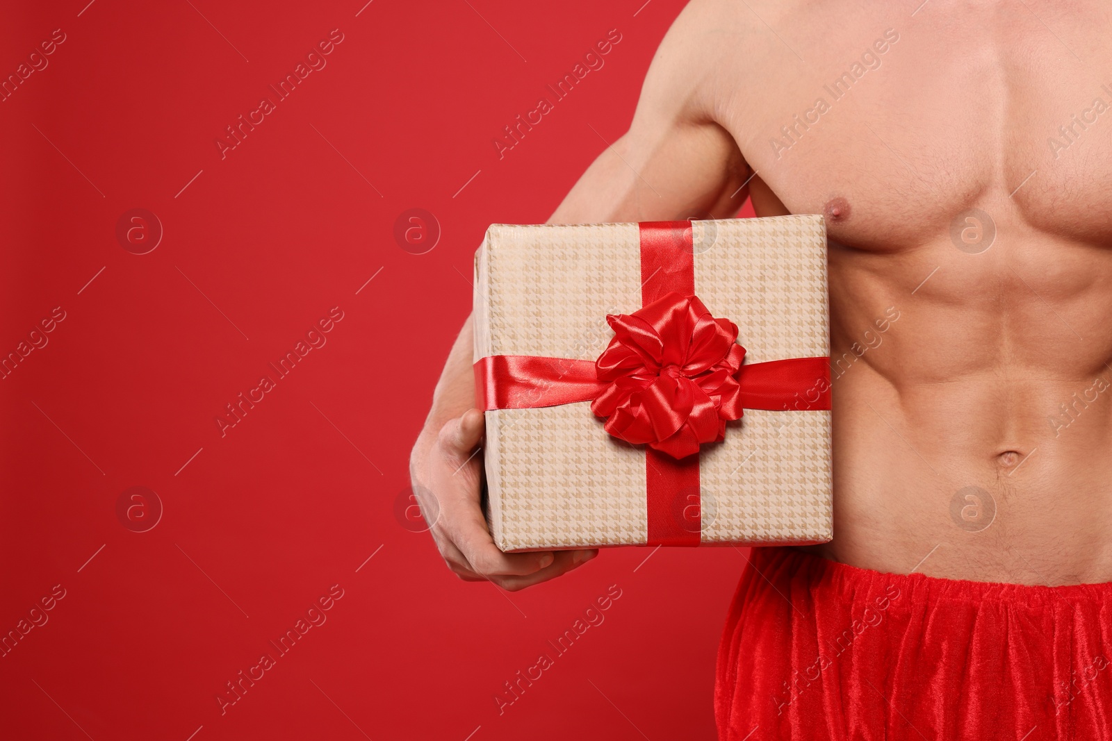 Photo of Young man with muscular body holding Christmas gift box on red background, closeup. Space for text