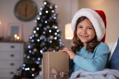 Photo of Cute little child in Santa hat with Christmas gift box sitting on sofa at home