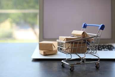 Internet shopping. Small cart with boxes near modern laptop on table indoors, space for text