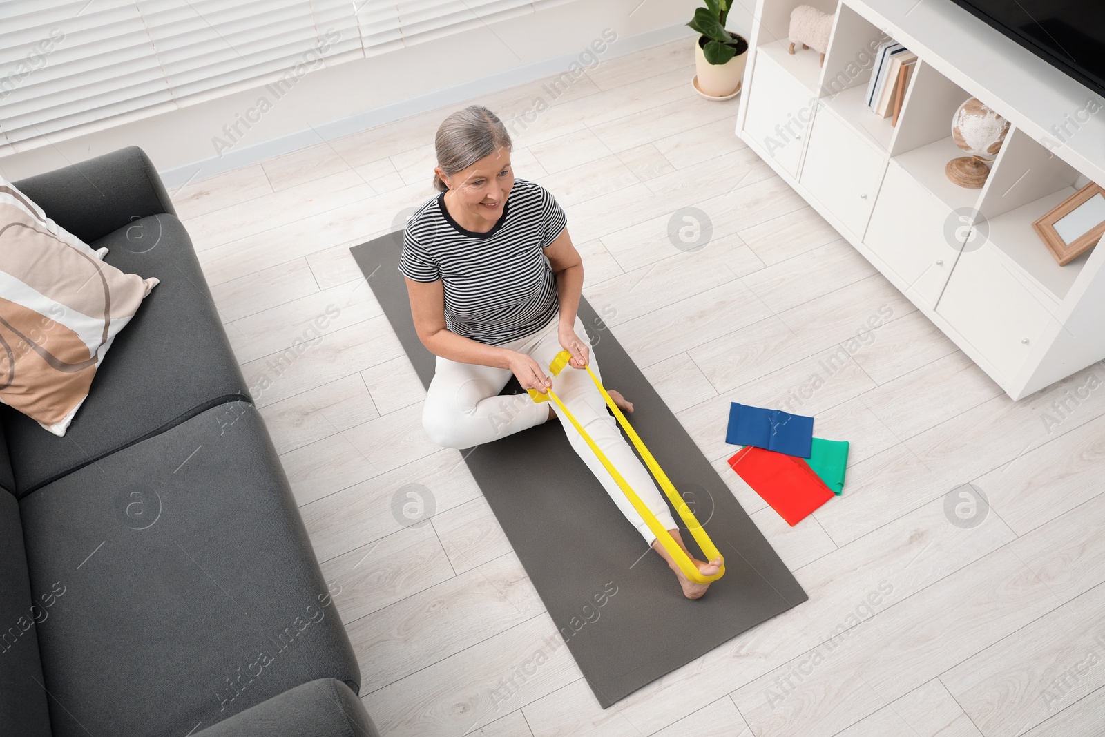 Photo of Senior woman doing exercise with fitness elastic band on mat at home, above view