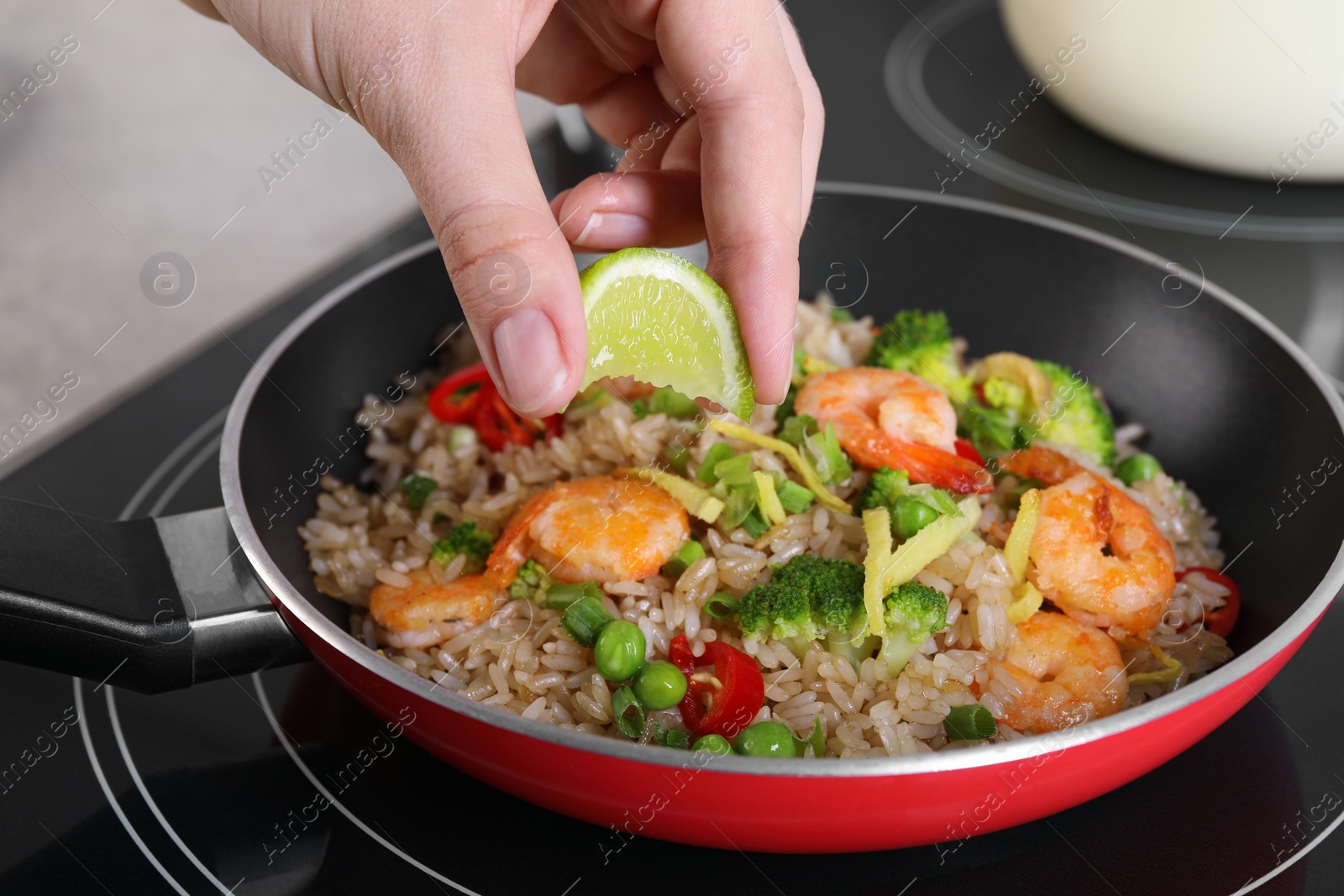 Photo of Woman squeezing lime into rice with shrimps and vegetables on induction stove, closeup