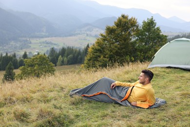 Photo of Man in sleeping bag near camping tent on hill, space for text
