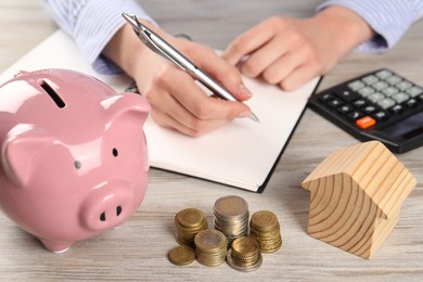 Woman planning budget at wooden table, focus on house model, coins and piggy bank