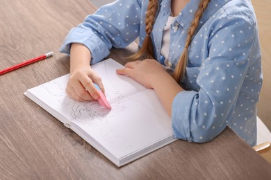 Photo of Girl erasing drawing in her book at wooden table, closeup