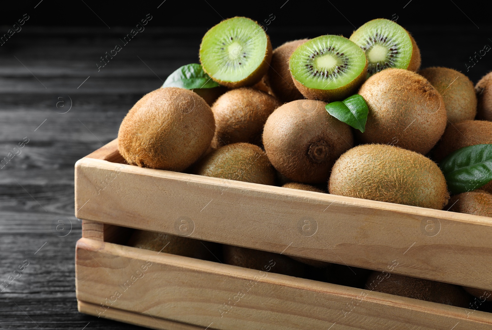 Photo of Crate with cut and whole fresh kiwis on black wooden table, closeup