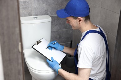 Photo of Young plumber writing results of examining toilet bowl in water closet