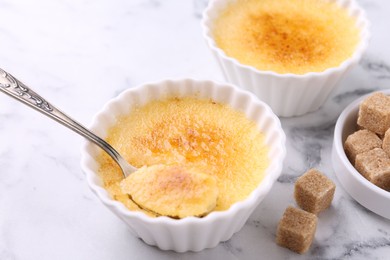 Photo of Delicious creme brulee in bowls, sugar cubes and spoon on white marble table, closeup
