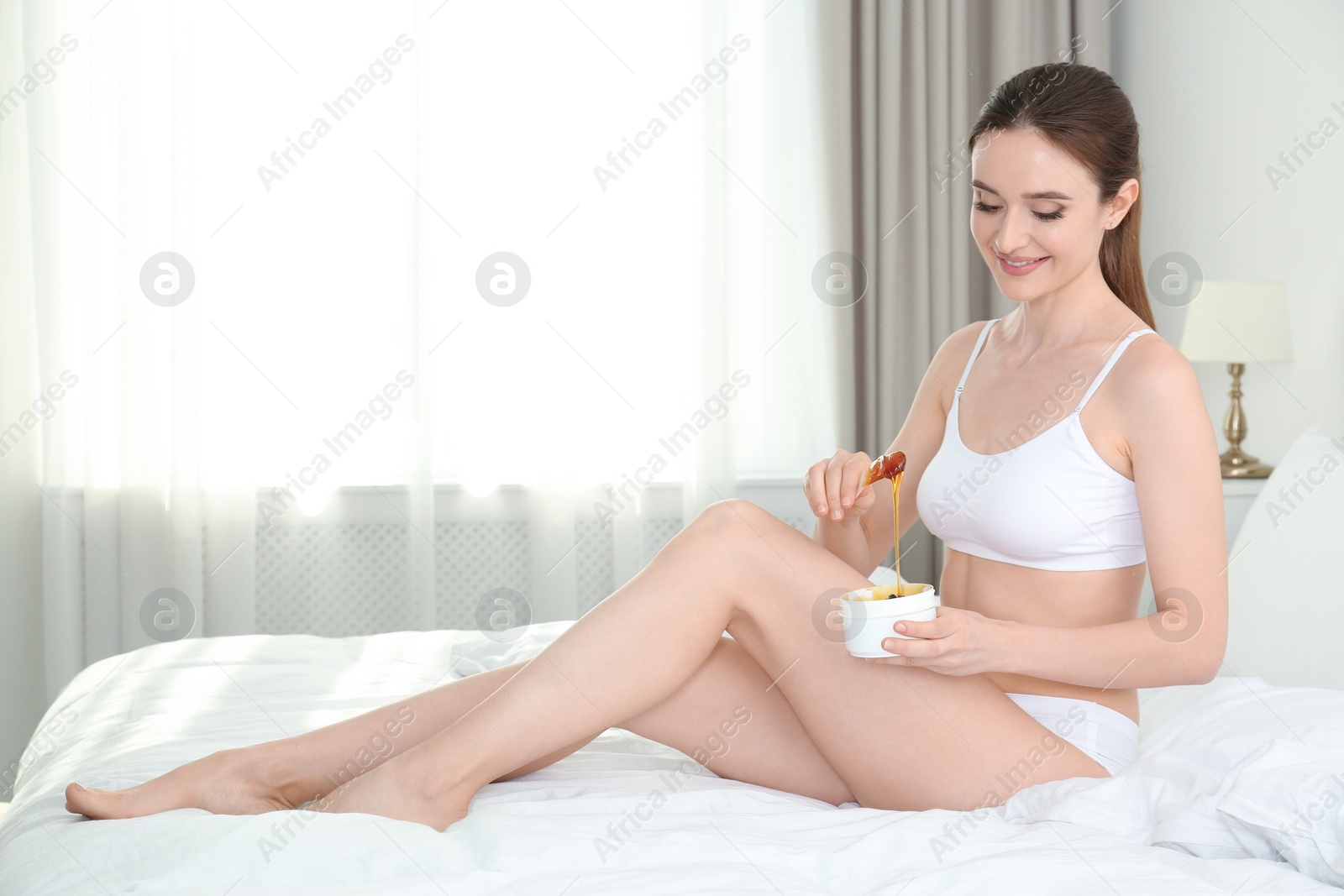 Photo of Smiling young woman holding hot wax for epilation procedure on bed at home. Space for text