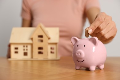 Woman putting money into piggy bank and holding house model at wooden table, focus on hand