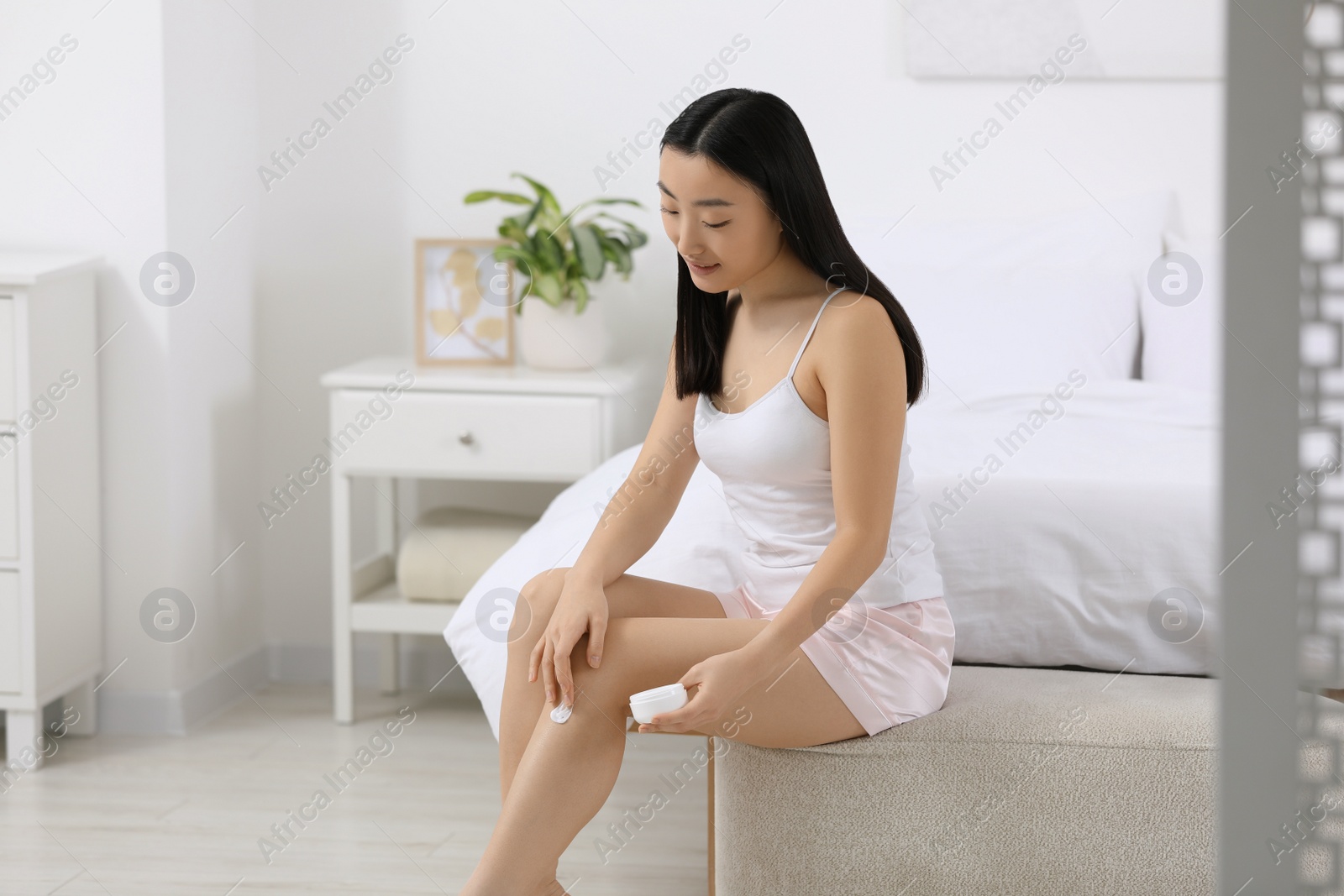 Photo of Beautiful young Asian woman applying body cream on leg in bedroom, space for text