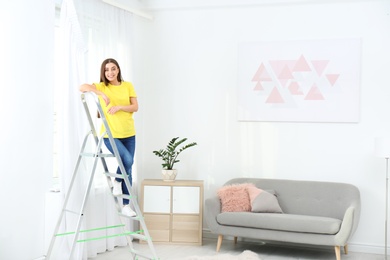 Young woman standing on stepladder near window with curtain in room. Space for text