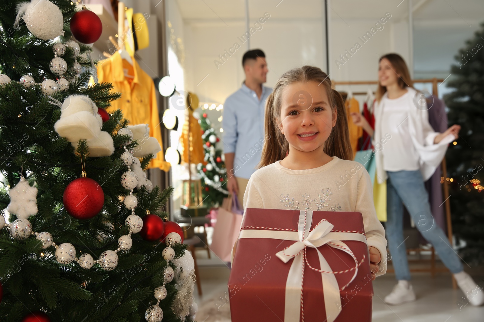 Photo of Little girl with gift box near her parents in store. Family Christmas shopping