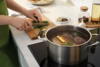 Photo of Homemade bouillon recipe. Woman cutting greenery in kitchen, focus on pot