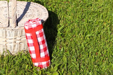 Rolled checkered tablecloth near picnic basket on green grass outdoors, space for text