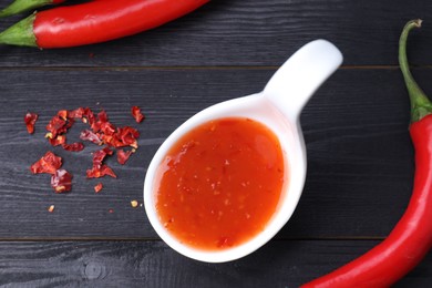 Photo of Spicy chili sauce in spoon and peppers on black wooden table, flat lay