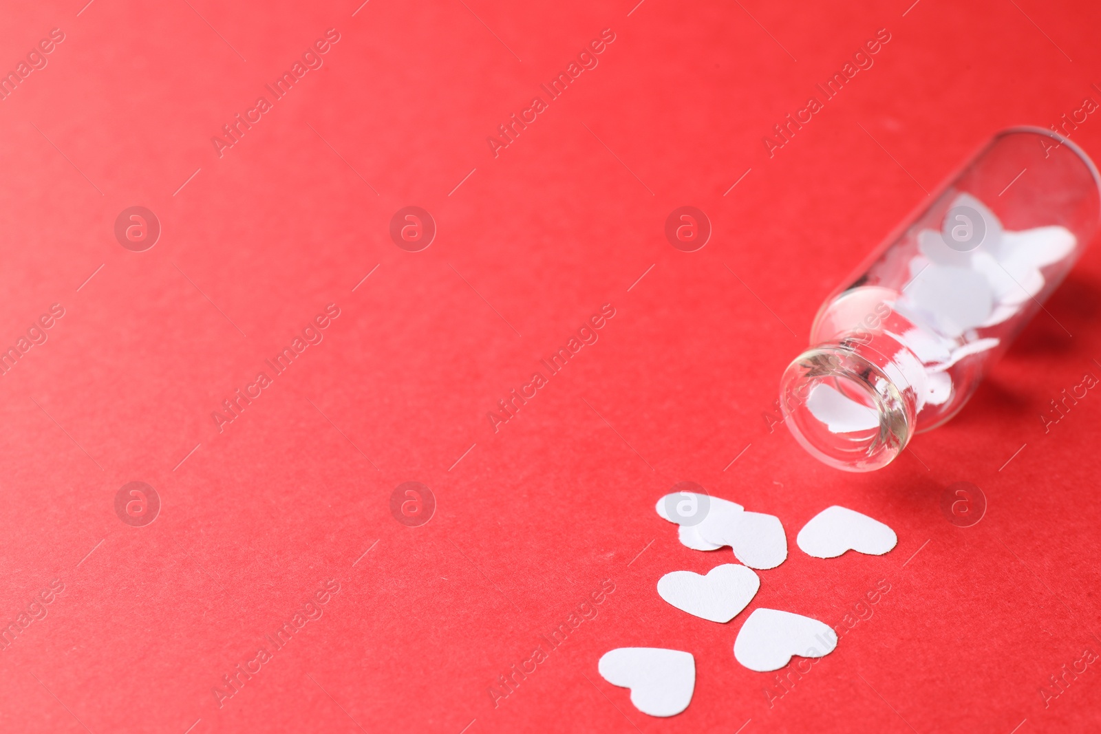 Photo of White paper hearts scattered from small glass bottle on red background. Space for text