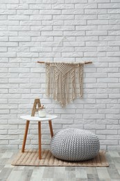 Comfortable knitted pouf, table and decor elements near white brick wall indoors. Interior design