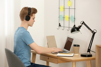 Photo of Teenage boy with headphones using laptop at wooden desk in room