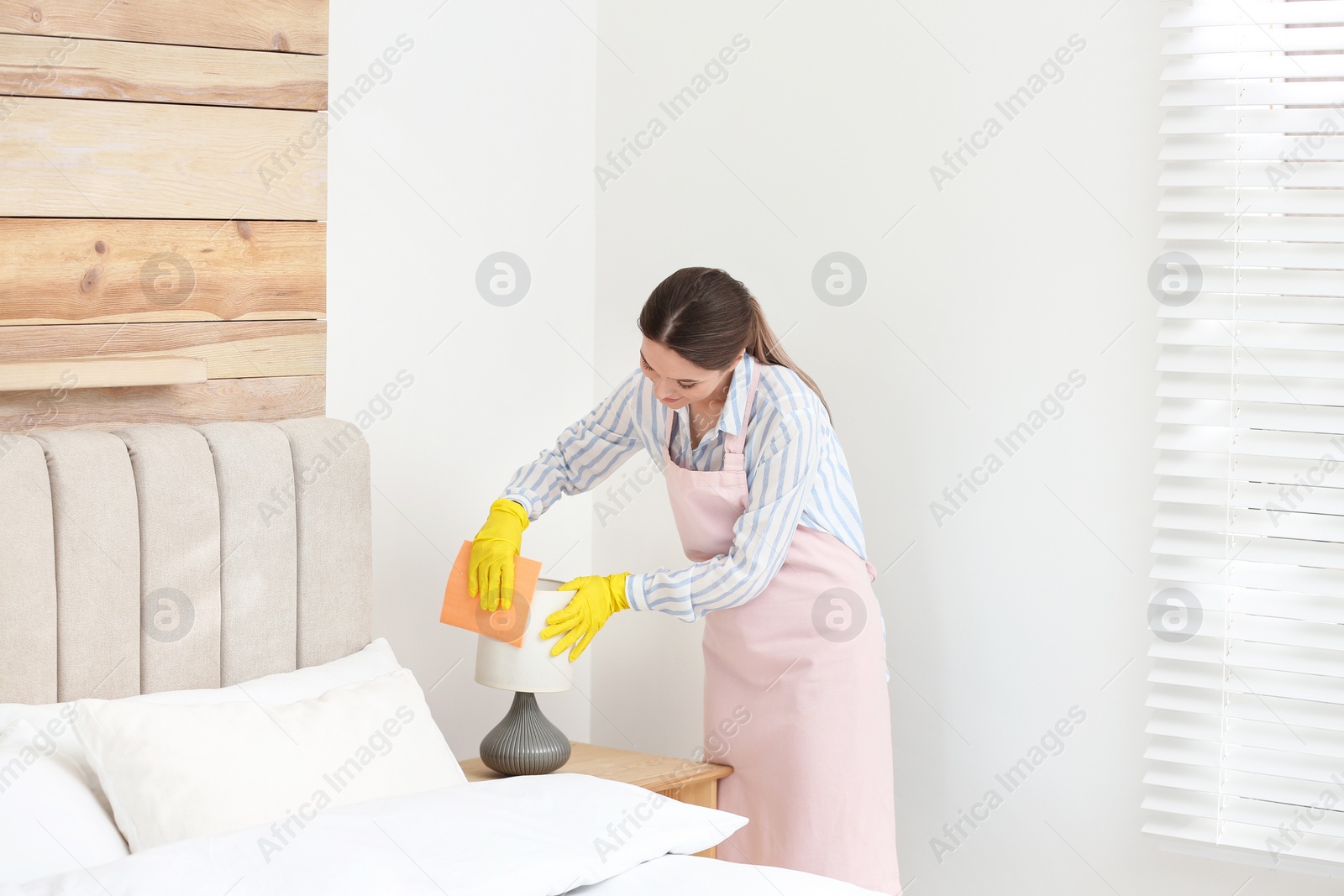 Photo of Young chambermaid wiping dust from lamp in bedroom