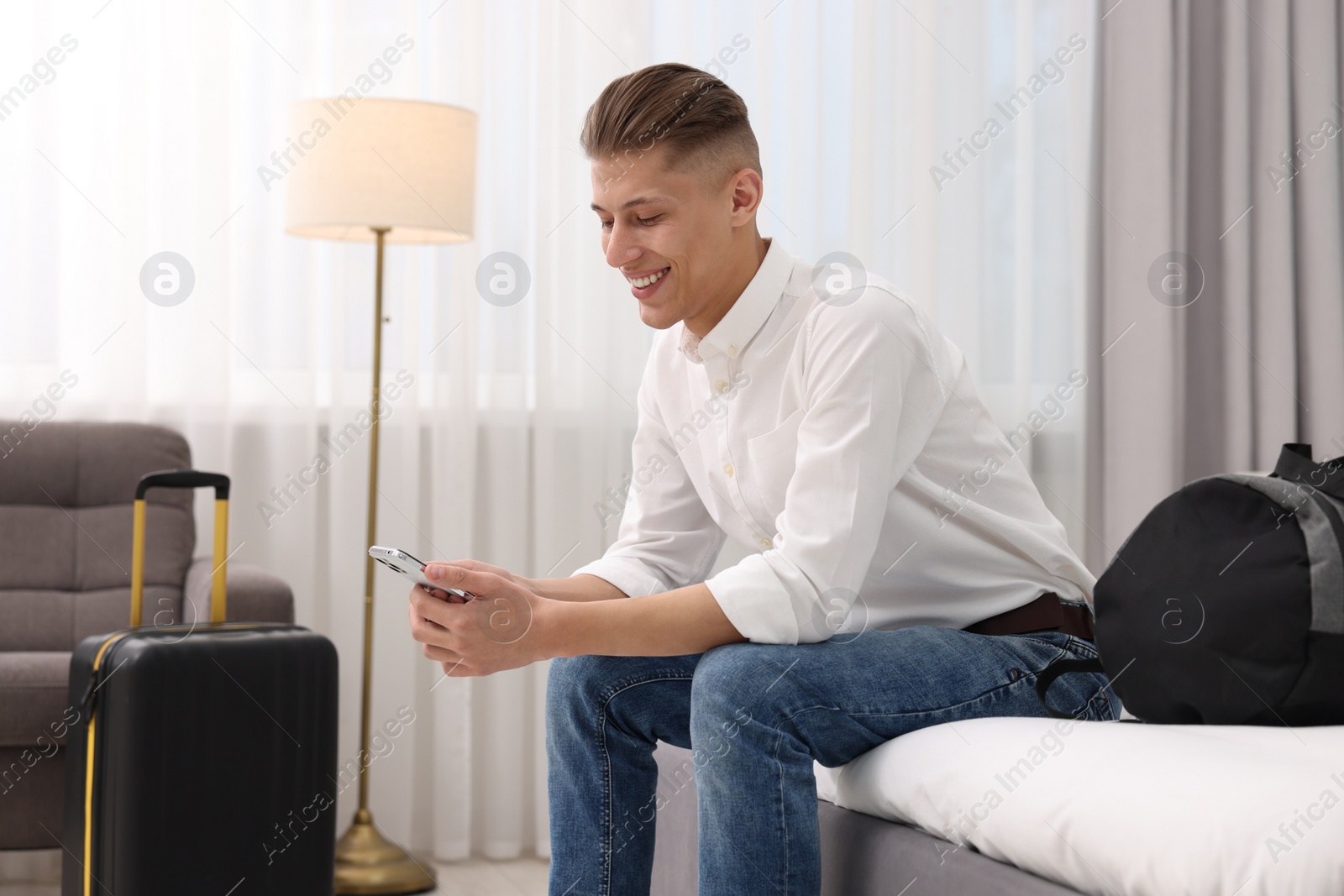 Photo of Smiling guest with smartphone relaxing on bed in stylish hotel room