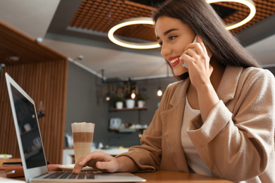 Photo of Young blogger with laptop talking on phone in cafe