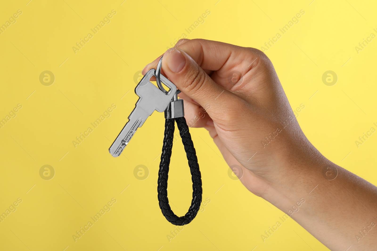 Photo of Woman holding key with black leather keychain on yellow background, closeup