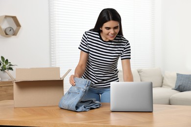 Photo of Young woman with just unpacked new jeans using laptop at table indoors. Online shopping