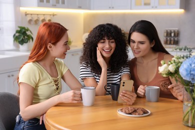 Happy young friends with smartphone spending time together at table in kitchen