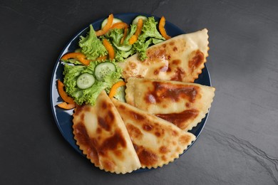 Photo of Delicious fried chebureki with vegetables served on black table, flat lay