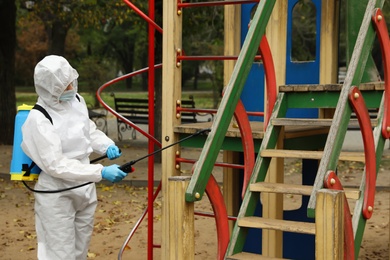 Photo of Woman wearing chemical protective suit with disinfectant sprayer on playground. Preventive measure during coronavirus pandemic