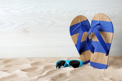 Stylish flip flops and sunglasses on sand near white wooden wall, space for text