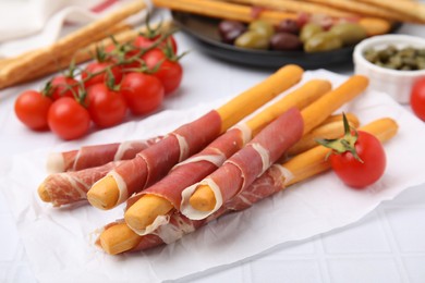 Delicious grissini sticks with prosciutto and snacks on white table, closeup