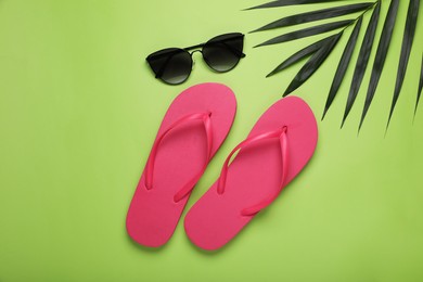Stylish pink flip flops, sunglasses and palm leaf on light green background, flat lay