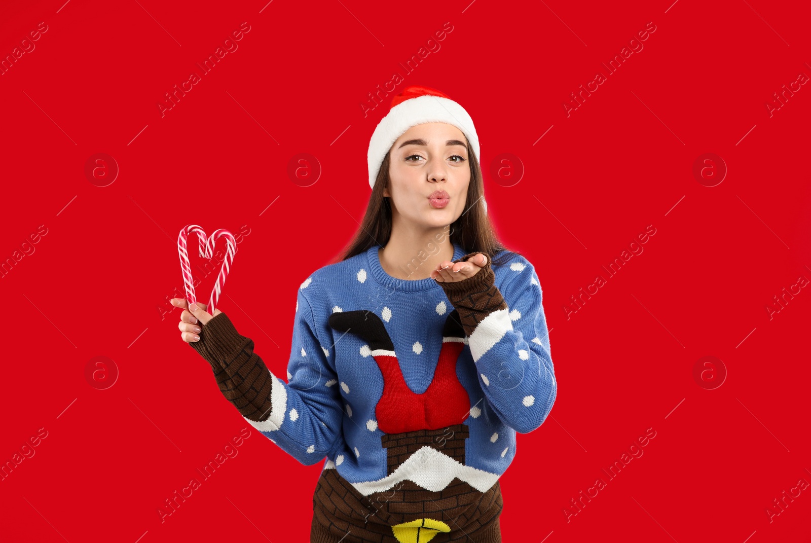 Photo of Young woman in Christmas sweater and Santa hat holding candy canes on red background