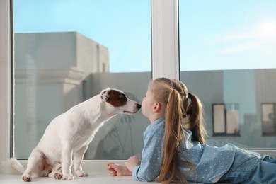 Photo of Cute little girl with her dog on window sill indoors. Childhood pet