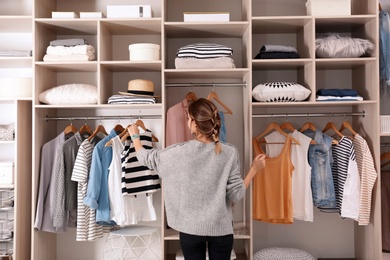 Photo of Woman choosing outfit from large wardrobe closet with stylish clothes and home stuff