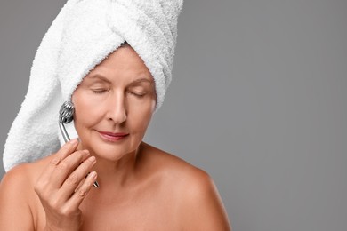 Woman massaging her face with metal roller on grey background, space for text