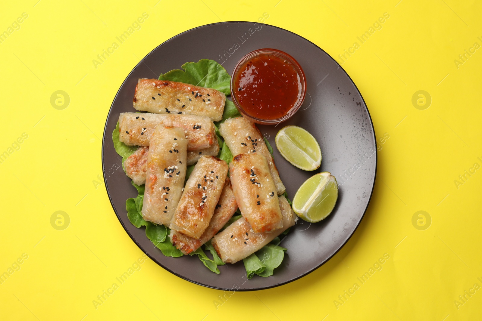 Photo of Tasty fried spring rolls with lettuce, lime and sauce on yellow table, top view