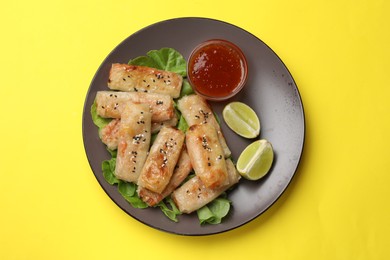 Photo of Tasty fried spring rolls with lettuce, lime and sauce on yellow table, top view