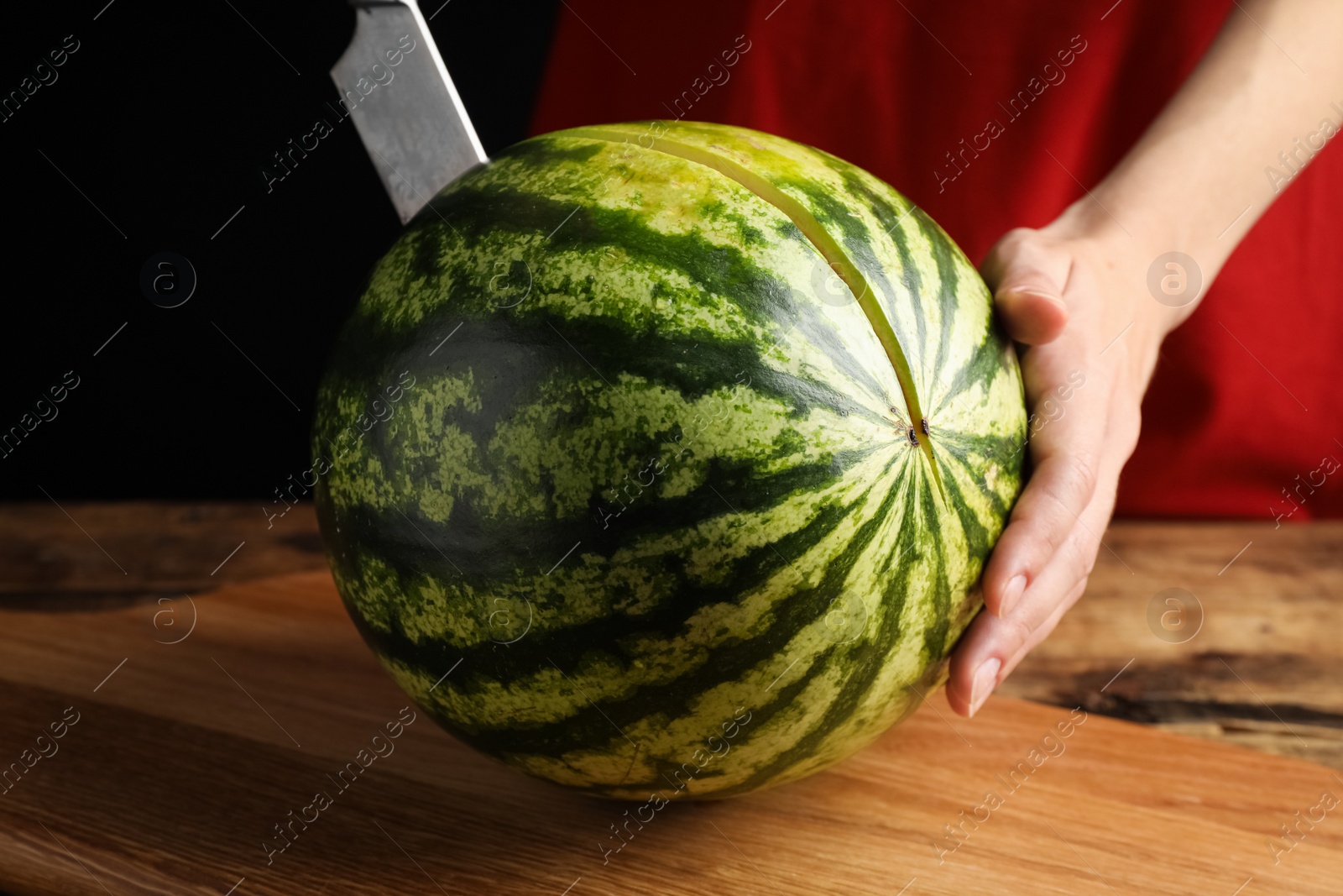 Photo of Woman cutting delicious watermelon at wooden table against black background, closeup