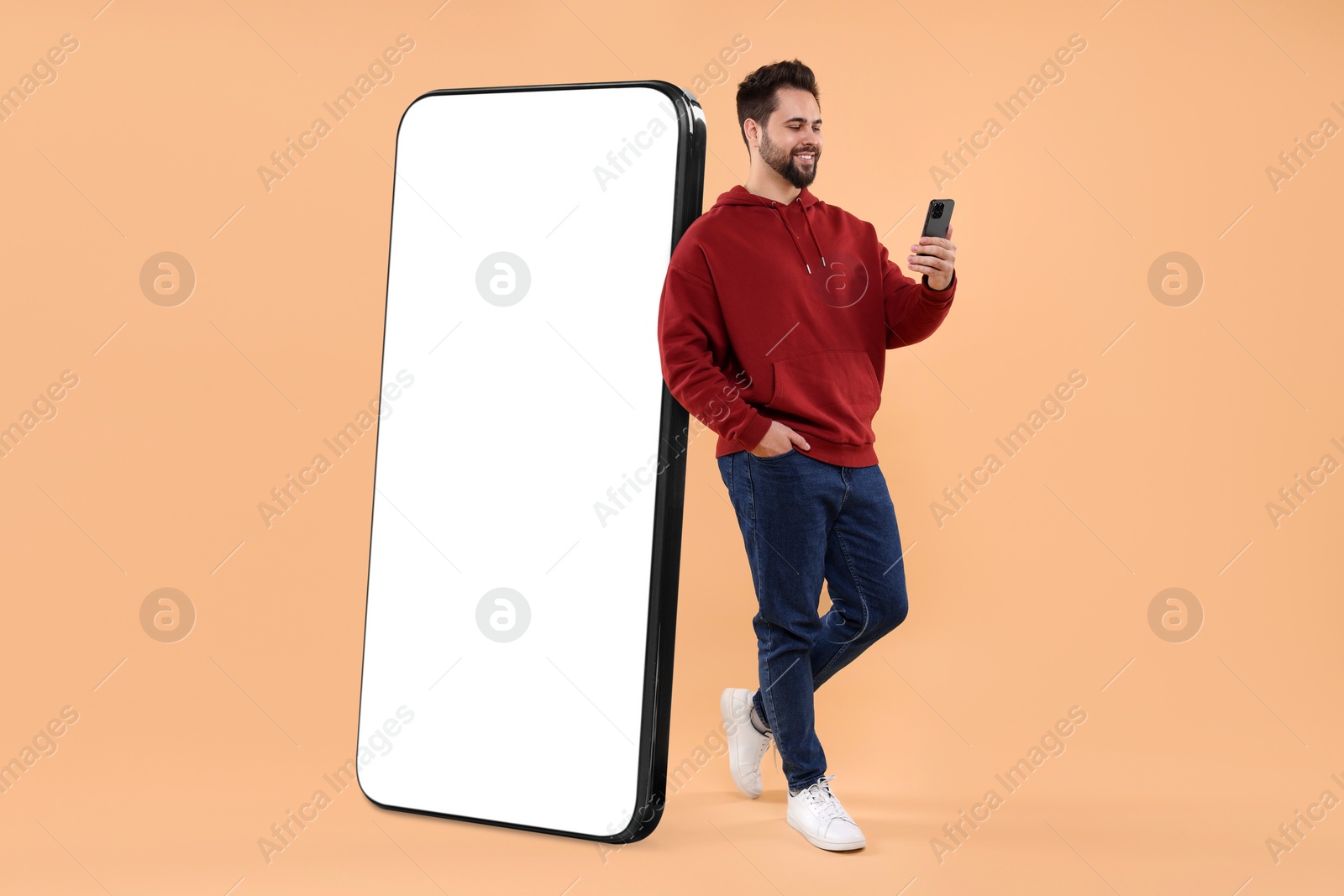 Image of Man with mobile phone standing near huge device with empty screen on dark beige background. Mockup for design