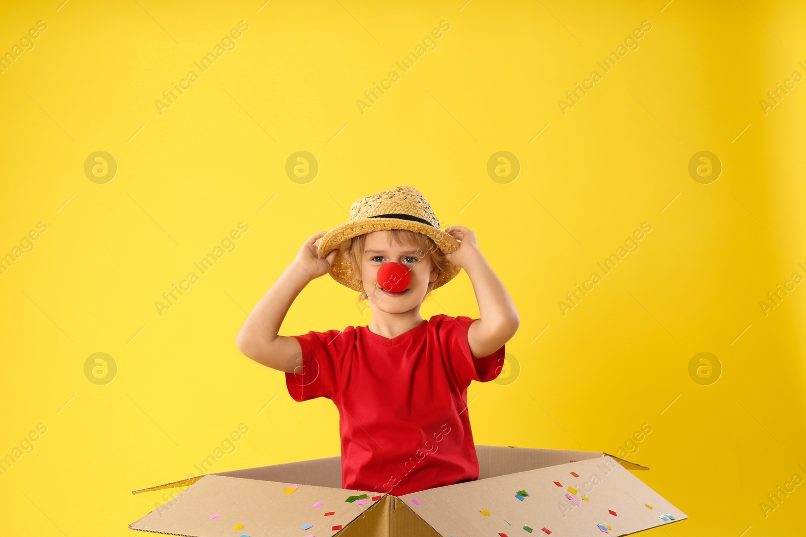 Photo of Funny little boy with clown nose in cardboard box on yellow background. April fool's day