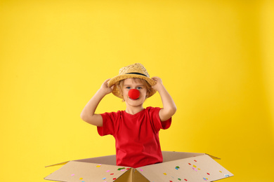 Photo of Funny little boy with clown nose in cardboard box on yellow background. April fool's day