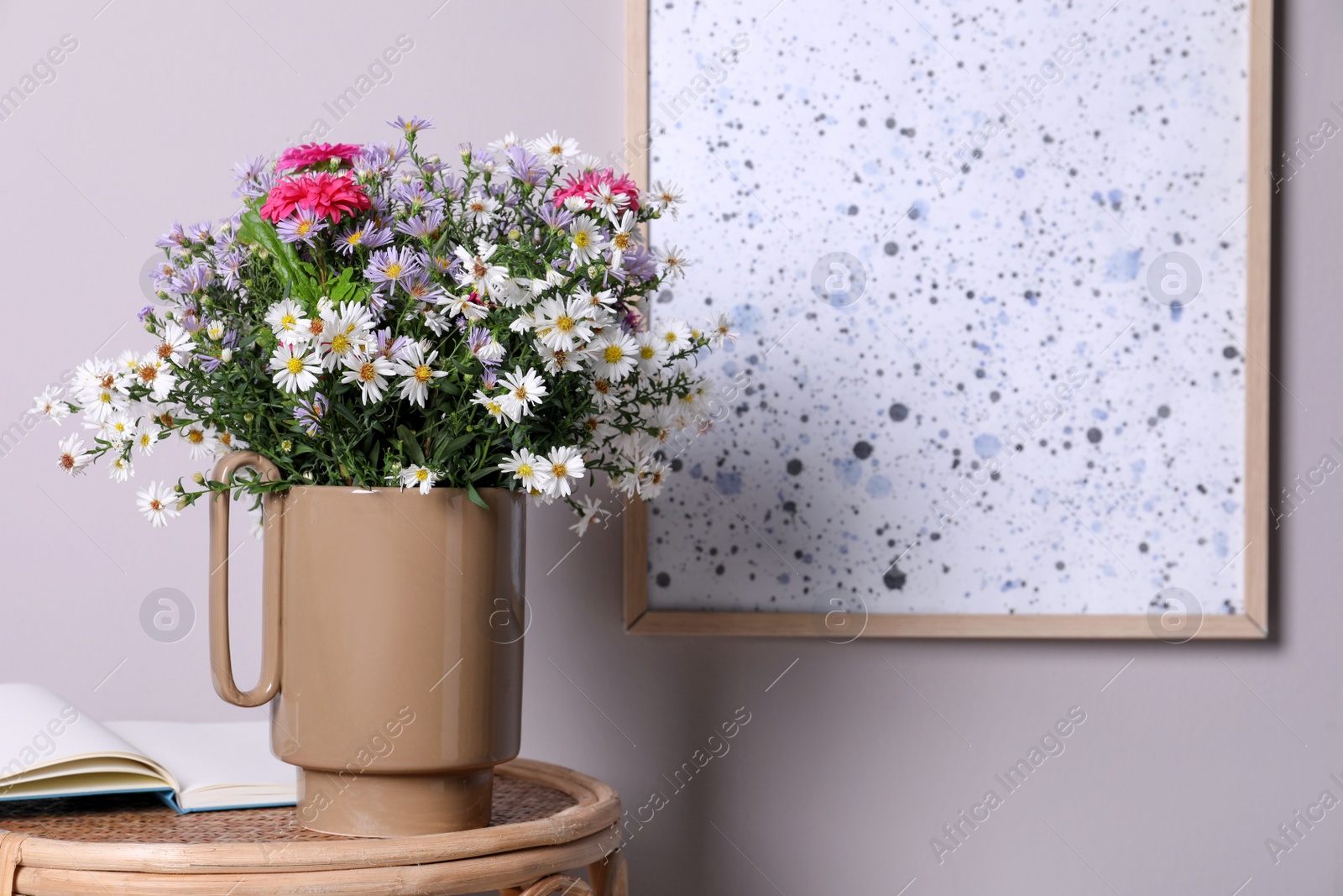 Photo of Stylish ceramic vase with beautiful flowers and open book on wicker table near light wall