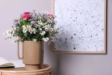 Stylish ceramic vase with beautiful flowers and open book on wicker table near light wall