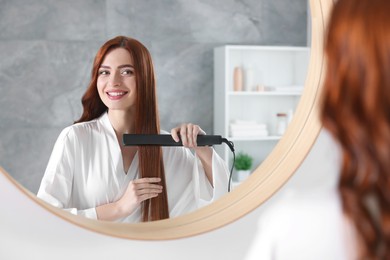 Photo of Beautiful woman using hair iron near mirror in room
