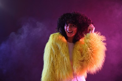 Photo of Beautiful young woman in yellow fur coat on color background in neon lights and smoke